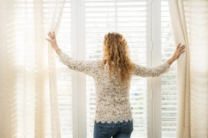 Woman looking out big bright window with draperies and blinds